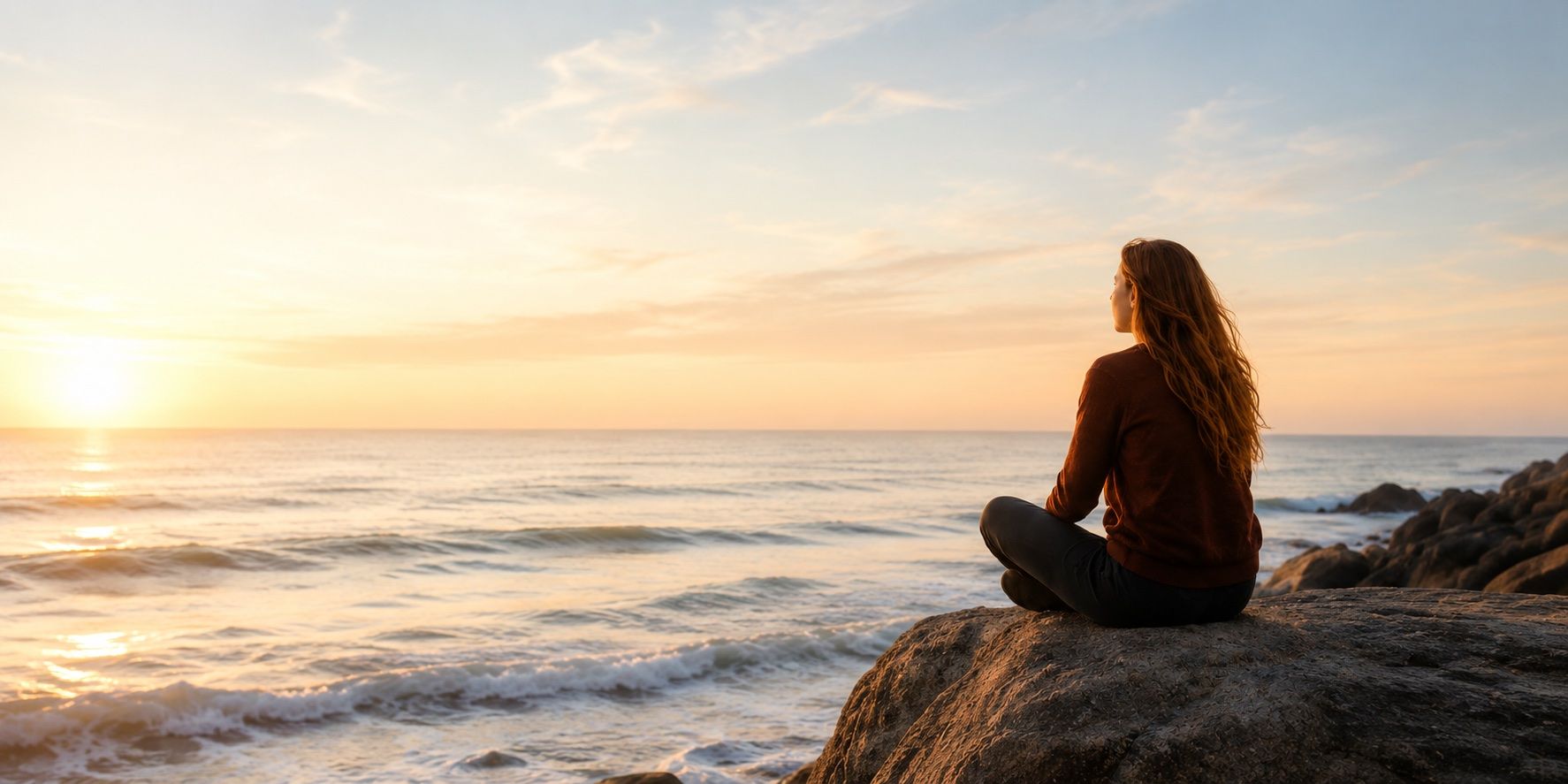 A woman in a warm sweater sitting on a coastal rock at sunset, looking out over the ocean.